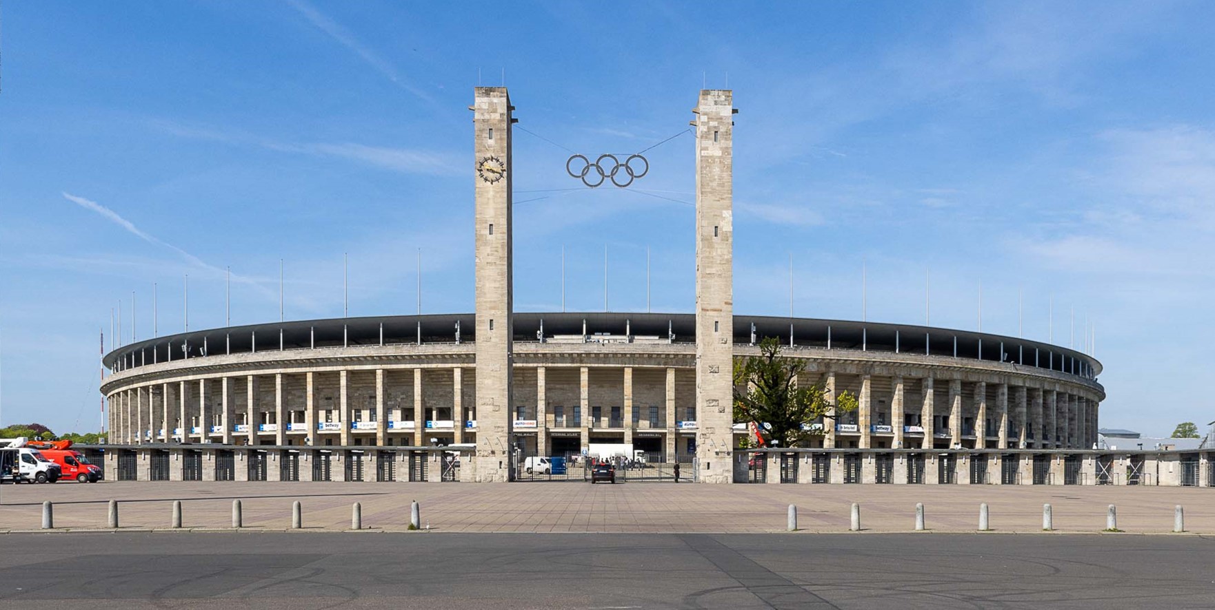 Ein Fußballstadion, davor zwei hohe Türme mit olympischen Ringen, der Himmel ist blau