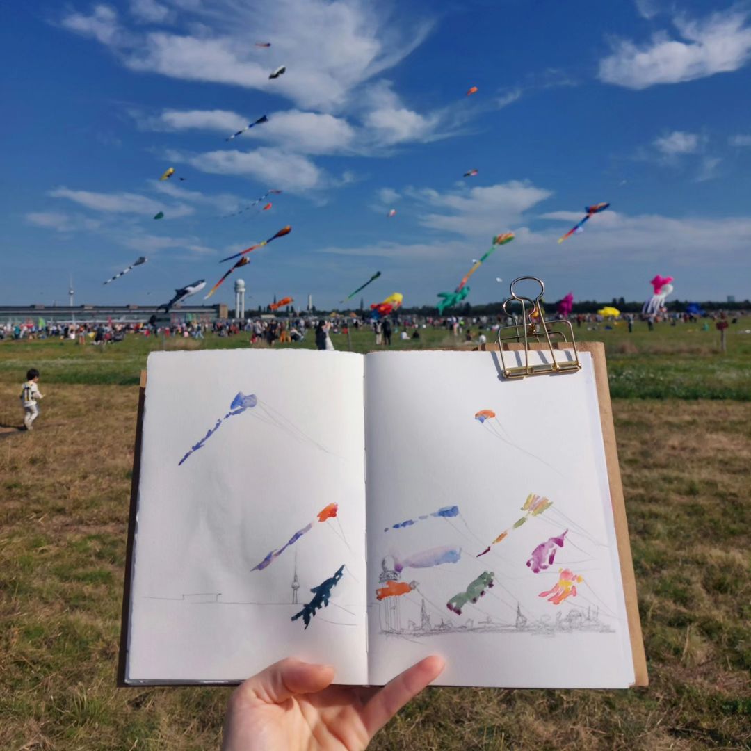 Skizzenbuch mit Riesendrachen, im Hintergrund das Tempelhofer Feld mit bunten Drachen